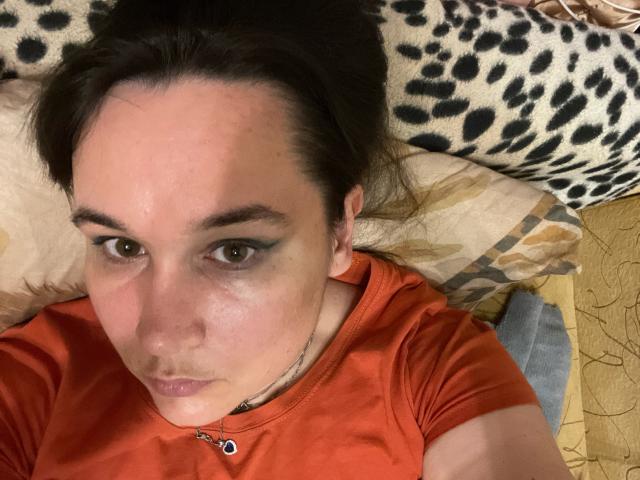 SaliBerry with green eyeshadow, brown hair, and a red shirt, lying on a pillow, showing a slightly annoyed face, and a small nose piercing.