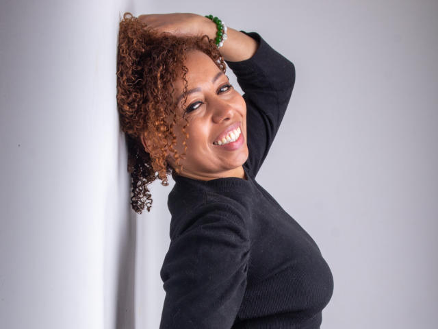 AryFoxy, com cabelos castanhos cacheados, sorrindo, vestindo uma blusa preta, tem a mão na cabeça, usa uma pulseira verde no pulso, mostrando os seios e um rosto bonito..