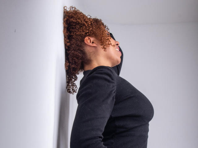 AryFoxy mit lockigem, braunem Haar, die ein schwarzes Langarmshirt trägt und an einer weißen Wand lehnt, blickt mit geschlossenen Augen nach oben und zeigt ihre kleinen, festen Brüste..
