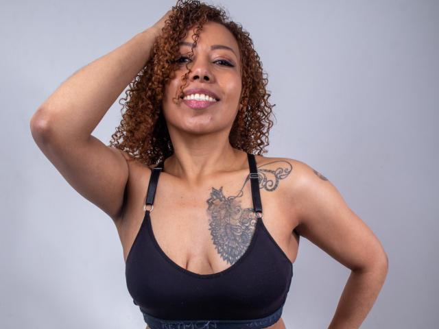 AryFoxy, con cabello castaño rizado, sonriendo, vistiendo un top deportivo negro, con una mano en el cabello, mostrando un tatuaje en el pecho y un tatuaje en el hombro izquierdo..