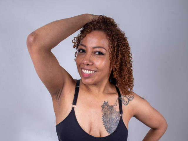 AryFoxy, com cabelo castanho encaracolado, sorrindo, usando uma regata preta, com o braço levantado, mostrando uma tatuagem no lado direito do peito, pele morena e seios pequenos, com um fundo cinza..