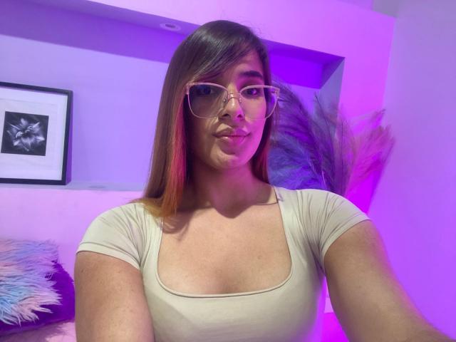 ValenSampaio with glasses, pouty lips, and small tits in a beige top, purple lighting, brown hair with red highlights, and a fluffy pillow in the background.