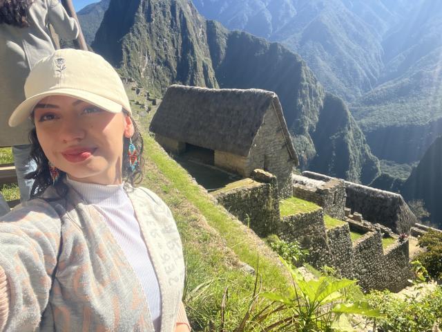 ElizaCanterville with a beige cap, colorful earrings, and a patterned cardigan, pouting with her pink lips, standing in front of a thatched roof building and green mountains.