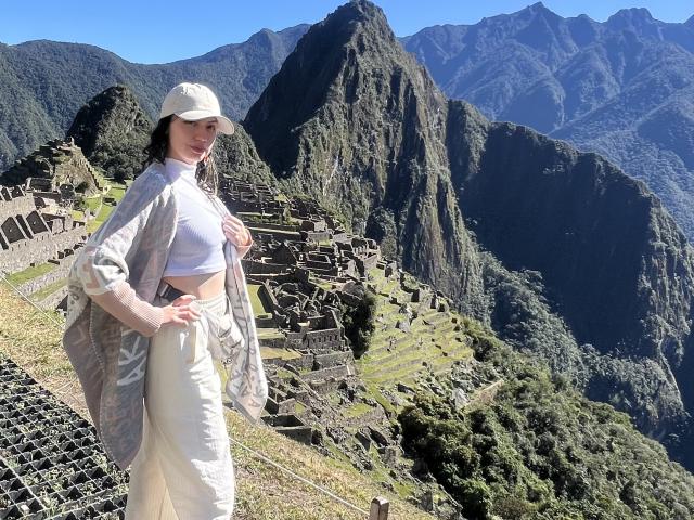 ElizaCanterville stands in front of Machu Picchu, wearing a white cap, open shirt, and beige pants, with her hand on her ass, showing her small tits.
