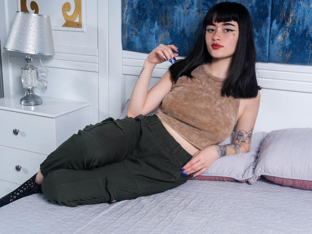 ScarlettBlaire com cabelo curto preto, regata marrom, calça verde, tatuagens, deitada na cama, olhando para a câmera, segurando um cigarro, unhas azuis, meias pretas, mesa de cabeceira branca, decoração azul e branca..