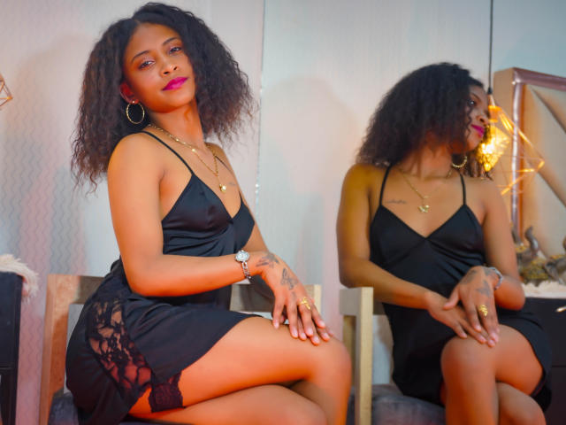 AntonellaiPetsch with curly hair, dark skin, and pink lipstick, wearing a black lace dress, sitting, reflected in a mirror, showing tattoos, gold jewelry, and a wristwatch.