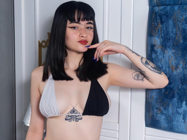 Scarlett Blaire mit schwartem Bob, einem weißen und schwarzen Bikini-Oberteil, blauen Nägeln, Tattoos und einem kleinen Brust-Tattoo, die in die Kamera schaut, mit einem Finger in der Nähe des Mundes. Sie hat helle Haut und trägt roten Lippenstift..