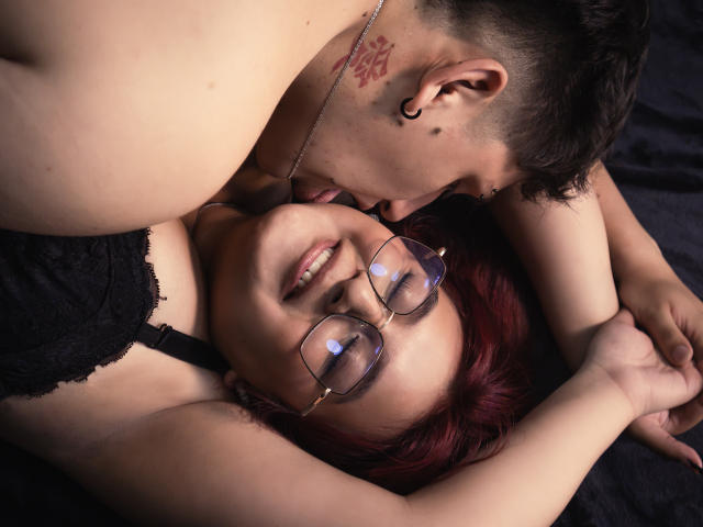 JaneAndLeo with red hair and glasses, laying down, tongue out, being licked by Leo with tattoos and short dark hair, wearing black lace bra, on dark fabric.