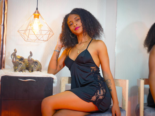 AntonellaiPetsch in a black satin dress with lace sides, sitting on a chair, curly hair, pink lipstick, gold hoop earrings, tattoo on right arm, looking at camera.