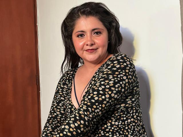 TiffanyAmazing, una donna robusta con capelli scuri e ondulati, indossa un abito nero con fiori, sorride leggermente e sta in piedi contro un muro bianco con una porta di legno sulla sinistra..