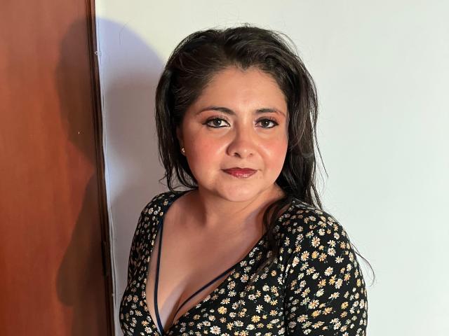 TiffanyAmazing with wet dark hair, wearing a black floral dress, showing her boobs in a deep V-neck, smiling with pink lipstick, standing against a white wall.