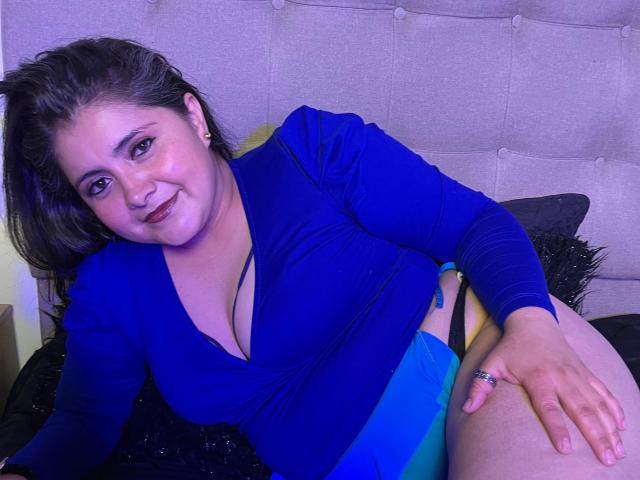 TiffanyAmazing with curvy ass, medium tits, and a blue V-neck top, sitting on a bed, smiling at the camera, wearing black underwear, and a ring on her right hand.
