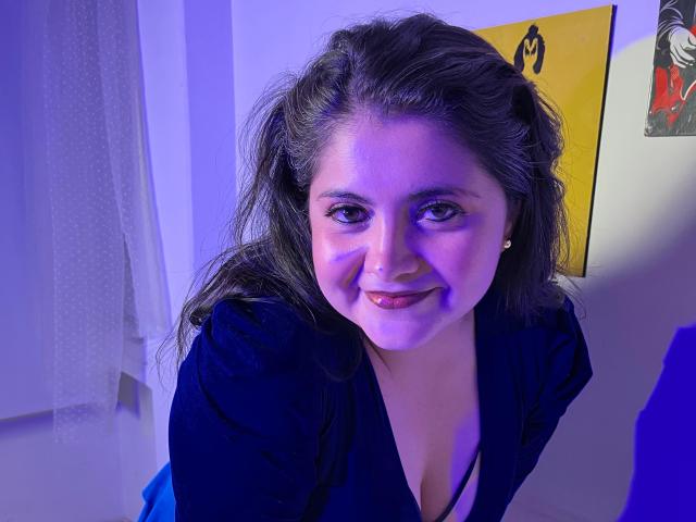 Tiffany, con i suoi capelli scuri e ondulati e un sorriso adorabile, indossa una maglietta nera con un taglio basso che mette in mostra il suo seno. È piegata in avanti, illuminata da luci blu e viola..