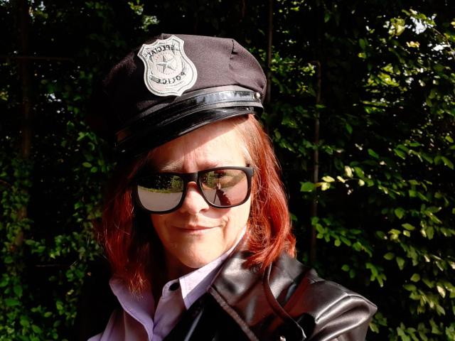 Kathy75-hot with red hair, wearing a black police cap, reflective sunglasses, and a leather jacket, standing in sunlight, showing a slight smile.
