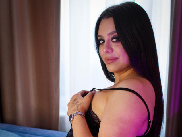 TaraBlues with long black hair, pinkish skin, and a tattoo on her right hand, wearing a black lace bra, looking back with a slight smile. Boobs visible.