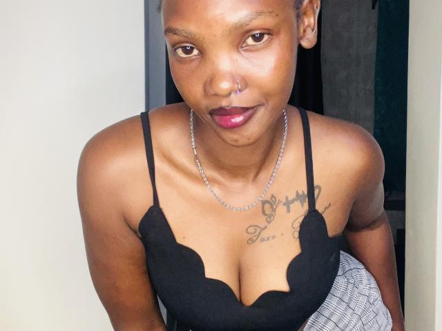 SexyMayana with dark skin, nose ring, and pink lipstick, wearing a black lace top, showing her tits, and a tattoo above her cleavage, in a silver chain necklace.