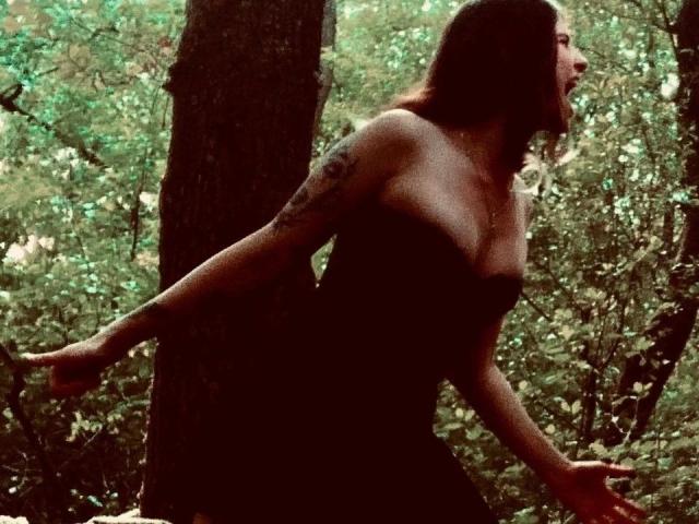 Silhouetted BloodyDiamond-hot with tits, arms out, leaning against a tree in a forest, wearing a dark dress, tattoo on right arm, brown hair, face turned to the side.