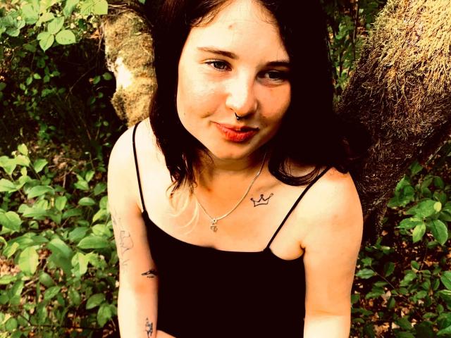 BloodyDiamond-hot with dark hair, nose ring, and tattoos, wearing a black spaghetti-strap top, standing in a forest, smiling with red lips, light skin, and a small cross necklace.