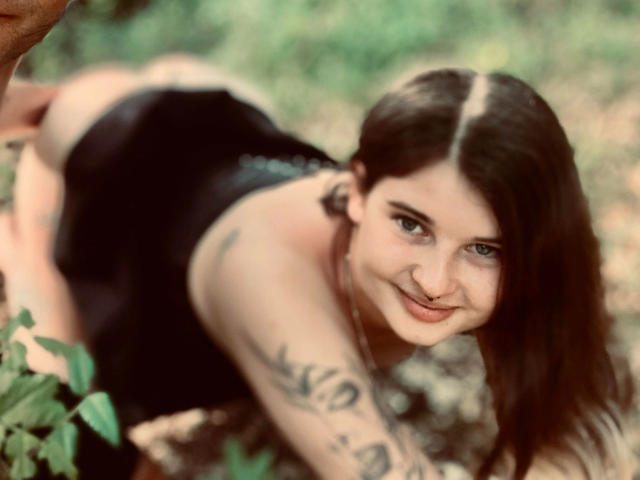BlodyDiamond-hot on all fours, smiling, with black lingerie, tattoos on her arm, and brown hair, looking at the camera, in a blurry outdoor setting.