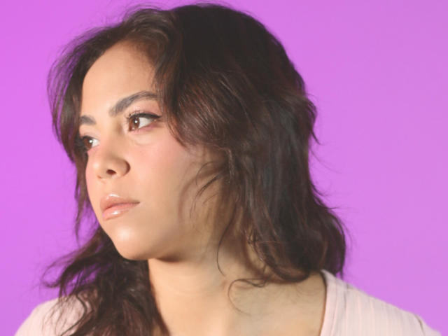 SamanthAdela heeft golvend donkerbruin haar, lichtbruine huid en volle lippen met een neutrale uitdrukking, en draagt een lichtroze top voor een paarse achtergrond..