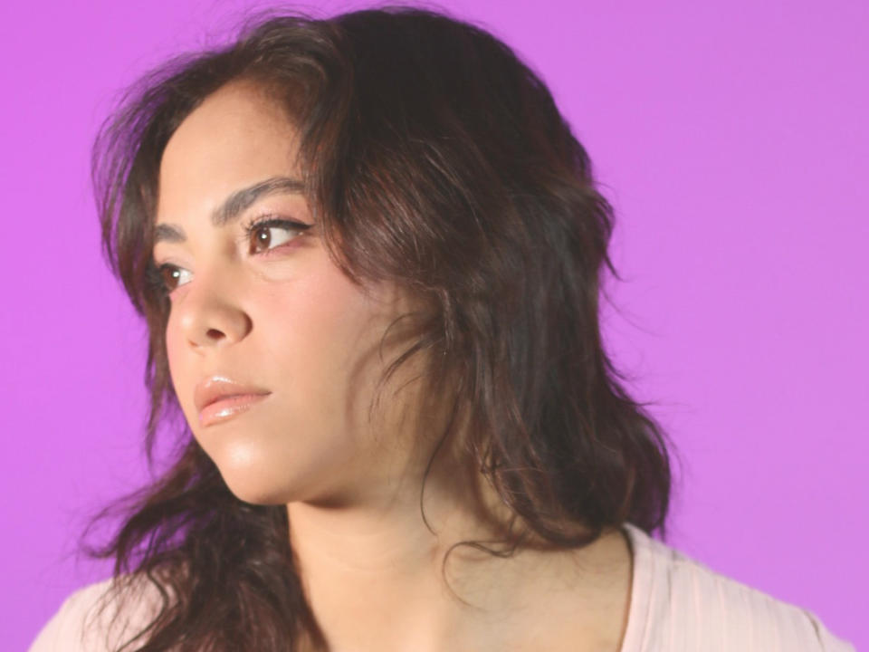 SamanthAdela tem cabelos castanho-escuros ondulados, pele clara e lábios cheios com uma expressão neutra, vestindo uma blusa rosa claro contra um fundo roxo..