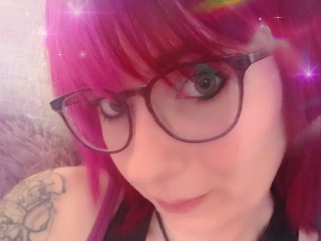 Ladyminniemaus-hot with pink hair, black glasses, and a tattoo on her shoulder, has a cute smile and green eyes. Sparkles are overlayed on the image.