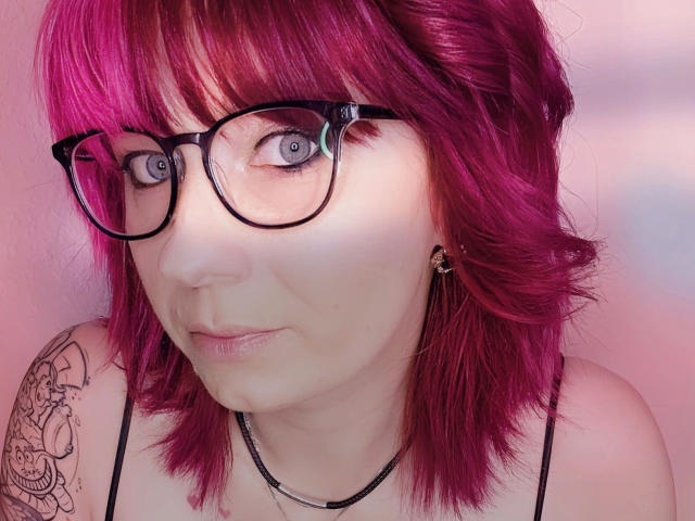 Ladyminniemaus-hot with pinkish-red hair, black glasses, and a tattoo on her left shoulder, wearing a black strap top, looking surprised with wide blue eyes.