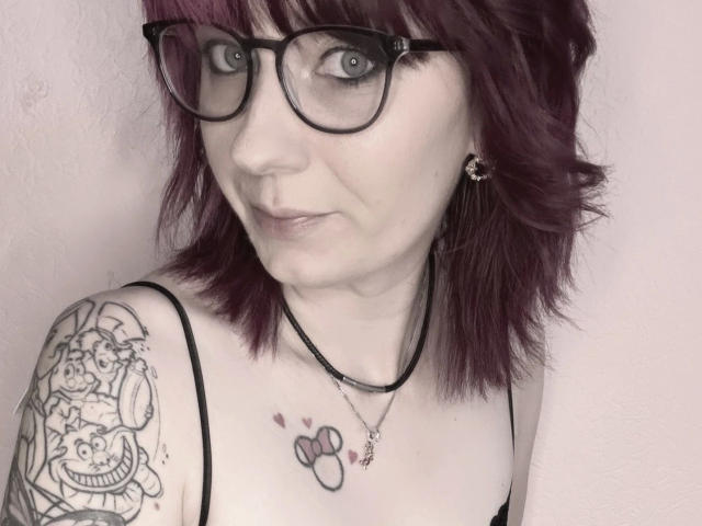 Ladyminniemaus-hot with purple hair, glasses, and tattoos, showing off her tits and a small Mickey Mouse tattoo on her chest. She's wearing a black spaghetti strap top.