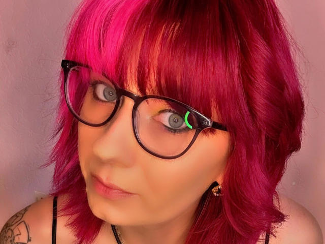 Ladyminniemaus-hot with bright pink hair, wearing black glasses, has a green light reflection in her eyes, and a tattoo on her left arm, looking slightly confused.