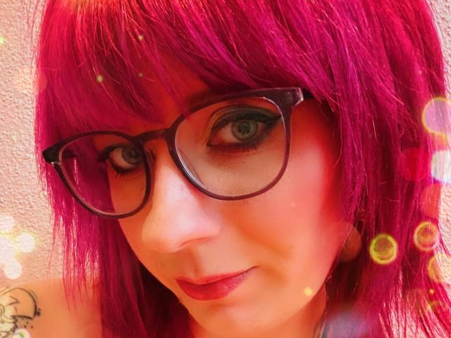 Ladyminniemaus-hot with bright pink hair, black glasses, blue eyes, and red lipstick, showing a slight smile, with subtle bokeh effects. Her ass tattoo is partially visible.