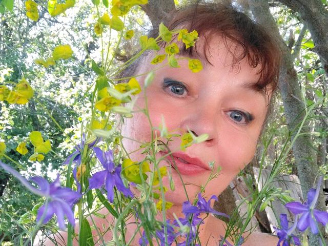 MilaMils with purple and yellow flowers in her face, blue eyes, pink lips, brown hair, slightly wavy, looking up, wearing a pink top, surrounded by greenery.