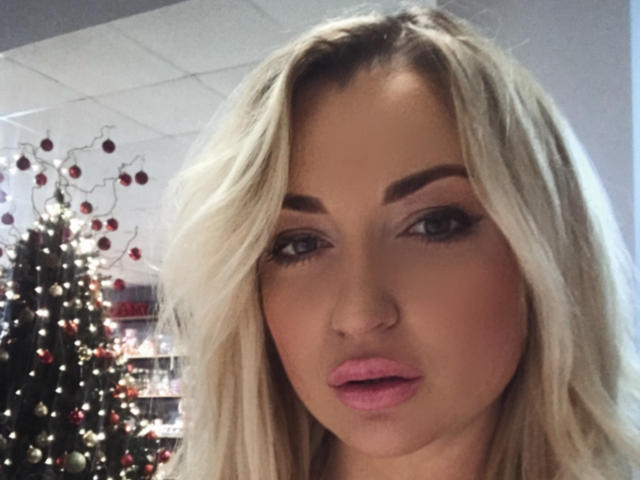 MillaLatvia with blonde hair, pink lips, and makeup, staring at the camera, showing her face and upper body, Christmas tree with lights in the background.