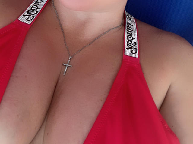 MillaLatvia's red halter top, showing her sizable boobs, with a silver cross necklace, and "PINK" straps visible. Her tan skin is slightly flushed.