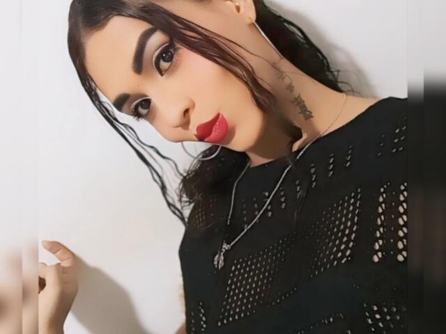 SexyHannaD con lápiz labial rojo, top de malla negra, tatuaje en el cuello, cabello oscuro en una coleta, y aros grandes, mirando a la cámara, sosteniendo su cabello a la izquierda..