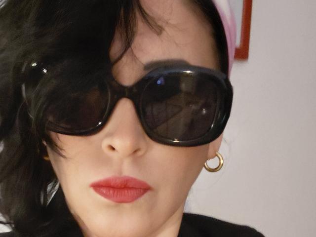 Claudias has dark wavy hair, black glossy sunglasses, red lipstick, and a gold hoop earring, looking serious with a slightly pouty expression.