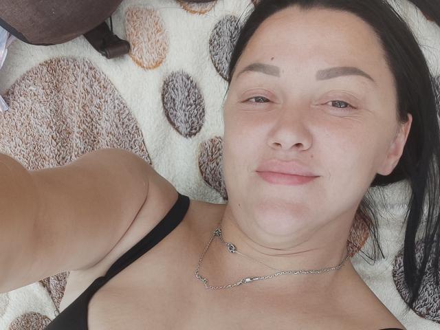 XCrazyBabeX lying on a patterned blanket, face up, black hair, brown eyebrows, no makeup, black tank top, silver necklace, slight smile, relaxed, fair skin, small tits.