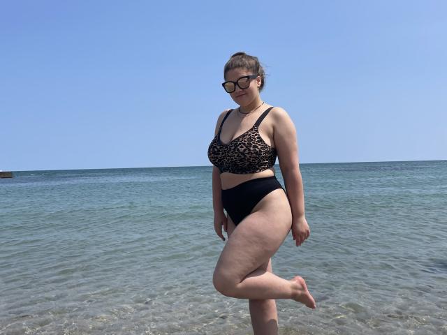 Madrinia, a plus-sized woman with pale skin and brown hair in a bun, wears a leopard-print bra and black bikini bottoms, standing in shallow blue water, leg raised.