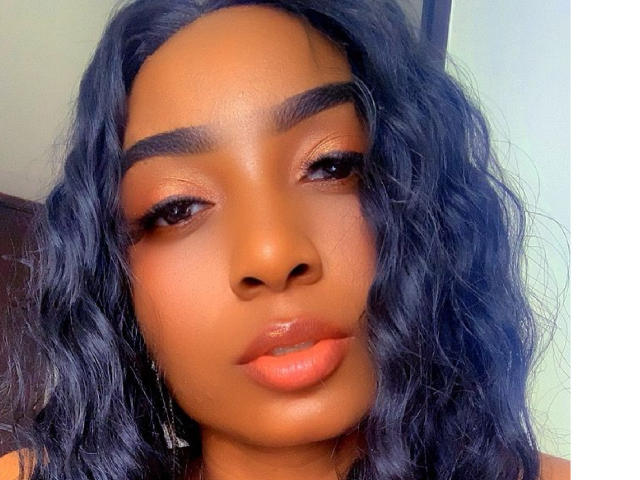 DIOSAPROHIBIDA has dark brown skin, wavy black hair, thick eyebrows, subtle makeup, and glossy orange lips. Her facial expression is neutral and slightly pouty.