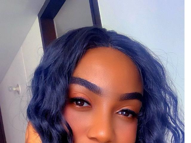 DIOSAPROHIBIDA has dark brown skin, curly black hair, thick eyebrows, and brown eyes with subtle eyeliner. Her face is centered in the image, showing a gentle smile.