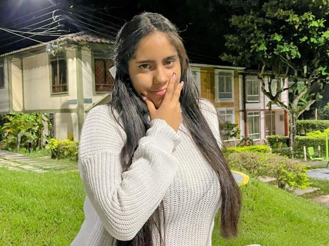 SiennaTaylor, con lunghi capelli neri, un maglione bianco lavorato a maglia, una mano sul viso, erba verde, una casa sullo sfondo, notte, che guarda la fotocamera, con un sorriso dolce, seno di medie dimensioni, carina..
