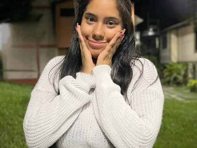 SiennaTaylor with hands on cheeks, smiling, wearing a white knit sweater, black hair, medium tits, pink nails, night outside, green grass, house in background, soft light.