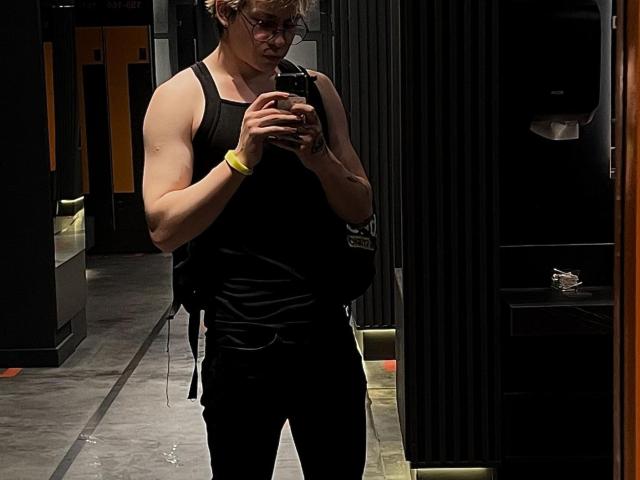 DesireCharlie with a muscular build, wearing a black tank top and black pants, standing in a dark hallway, looking at a phone, with a yellow bracelet on his wrist.