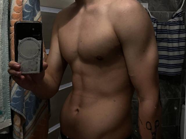 Shirtless DesireCharlie with a muscular, toned, and slightly hairy upper body, holding a phone in front of their face, showing a tattoo on their right arm and a visible navel.