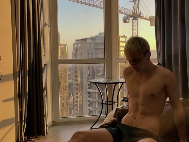 Shirtless DesireCharlie with blond hair and glasses, wearing green shorts, sitting on bed in dimly lit room, looking down, hands on knees, cityscape with cranes visible through window.