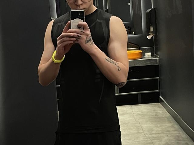 DesireCharlie, sleeveless black top, muscular arms, tattoos, yellow bracelet, holding phone, taking selfie, standing in dark room, visible ass, short black skirt, glasses, phone case.
