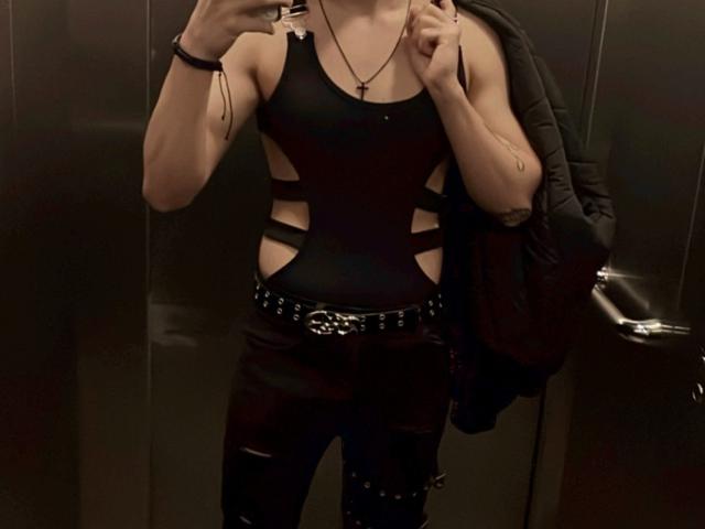 DesireCharlie in a black, cut-out tank top with side straps, black pants with metal studs, and a black belt, holding a black jacket, in a dimly lit room.