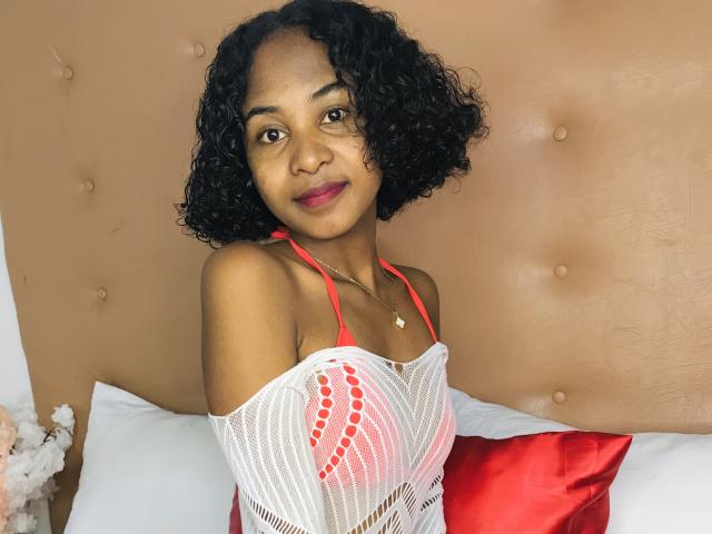 HotPoupee69 com cabelo preto encaracolado, pele morena, top de alça vermelha, tecido de malha branca cobrindo os seios, travesseiro vermelho, cabeceira bege, sorrindo, batom vermelho, colar, ambiente de quarto..