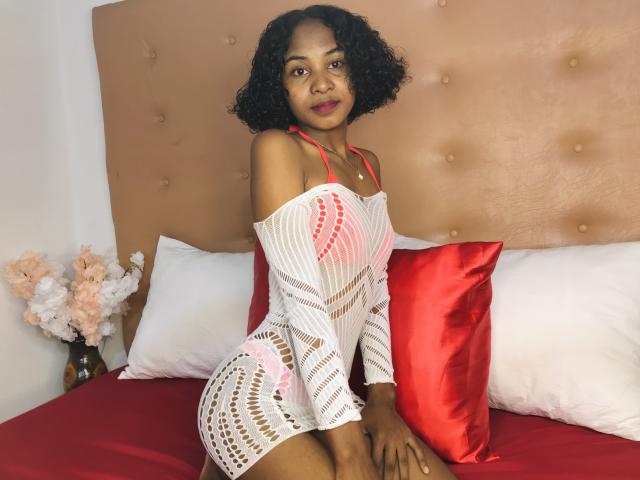 HotPoupee69 with curly black hair, wearing a white, off-shoulder, mesh dress, showing her ass, on a bed with red pillows, and a beige tufted headboard.