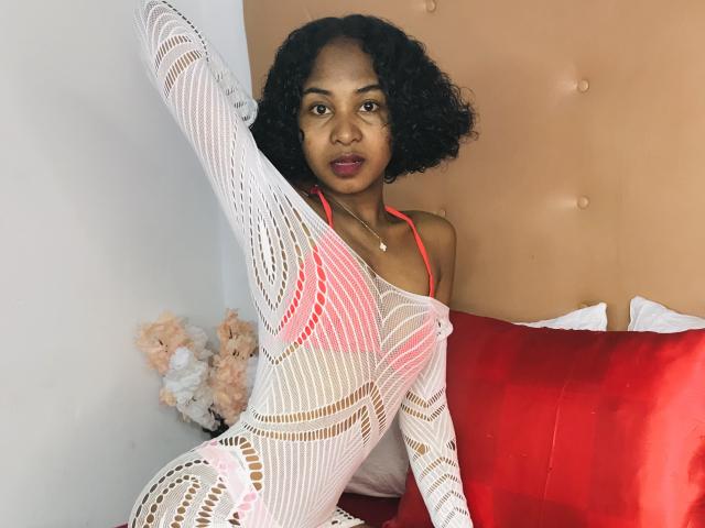 HotPoupee69 with curly black hair, medium brown skin, wearing a white, see-through, fishnet bodysuit, raising one leg, showing her pussy through the fabric, sitting on a red pillow.