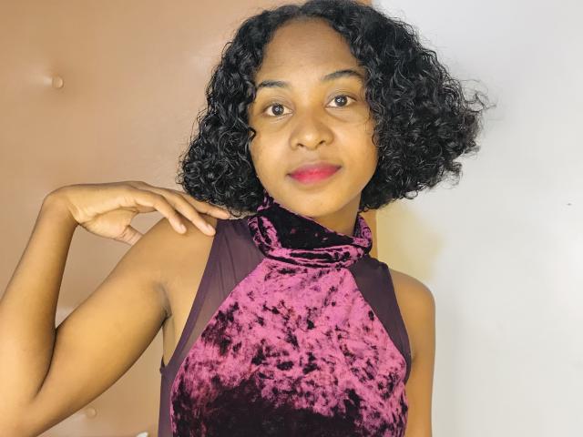 HotPoupee69 com cabelo preto encaracolado, pele morena e batom vermelho, vestindo uma blusa de alça roxa de veludo, com a mão no ombro, sorrindo. Seios visíveis sob a blusa, de tamanho médio..